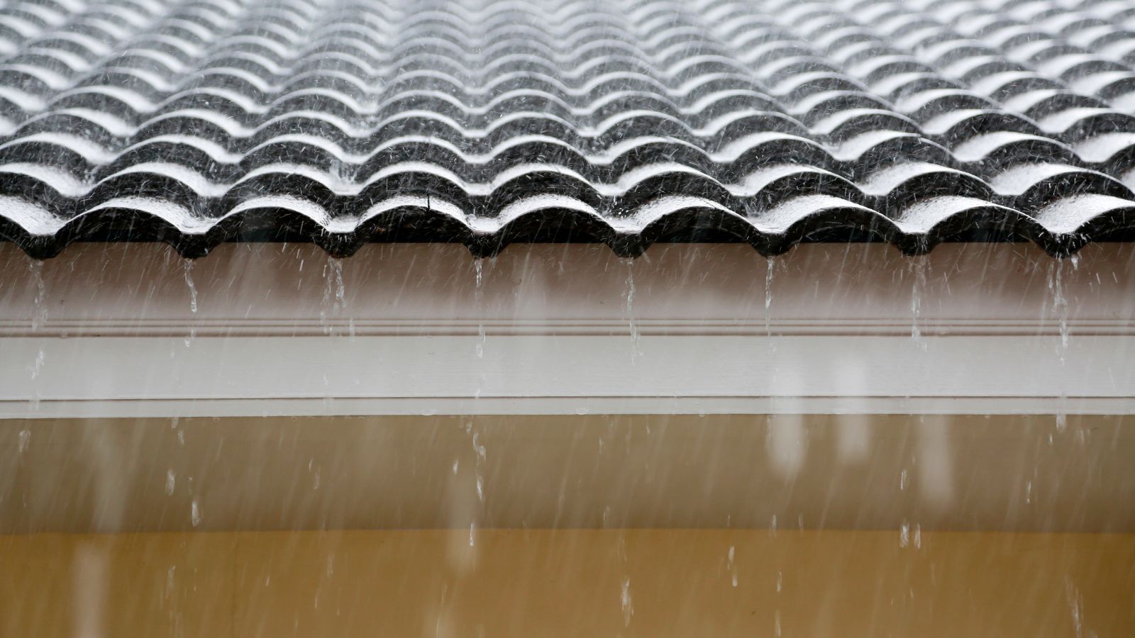 Lluvia sobre un tejado de tejas oscuras, con agua que cae por el borde sobre una franja blanca y una pared amarilla.